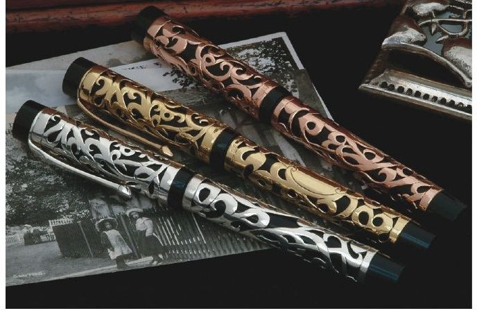 Three uncapped fountain pens side by side with identical pierced foliate metal overlays over black barrels, differing only in the colour of the metal.